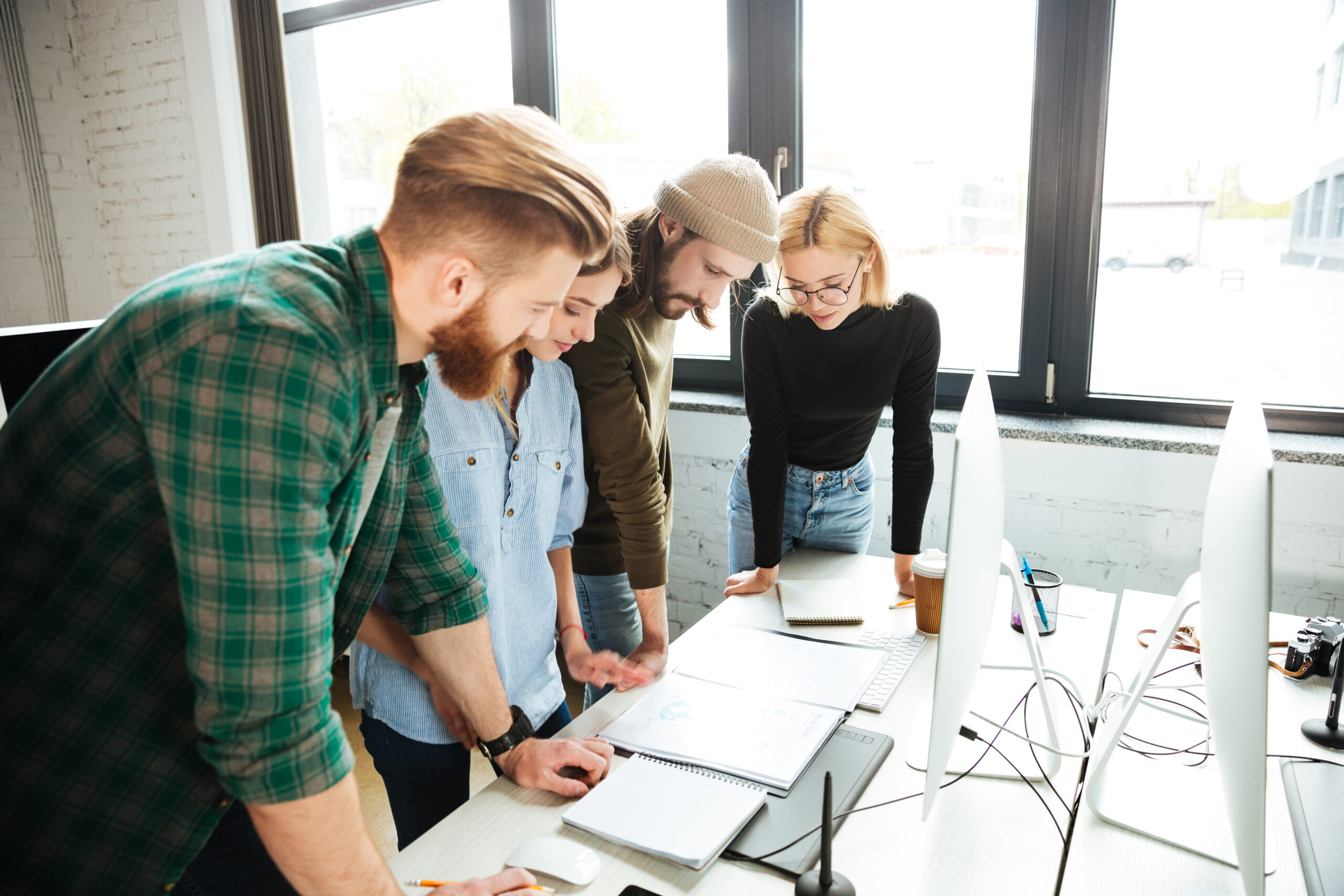 Photo of young concentrated colleagues standing in office using computer and documents. Looking aside.